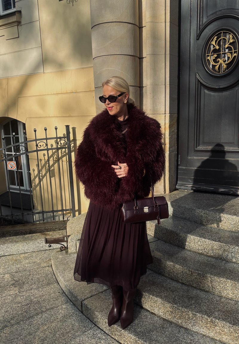 Burgunderfarbener Kunstpelzmantel, schwarzes Kleid, passende knielange Stiefel und Handtasche. Ergänzt durch Sonnenbrille und silbernen Schmuck, vor einer Steintreppe.