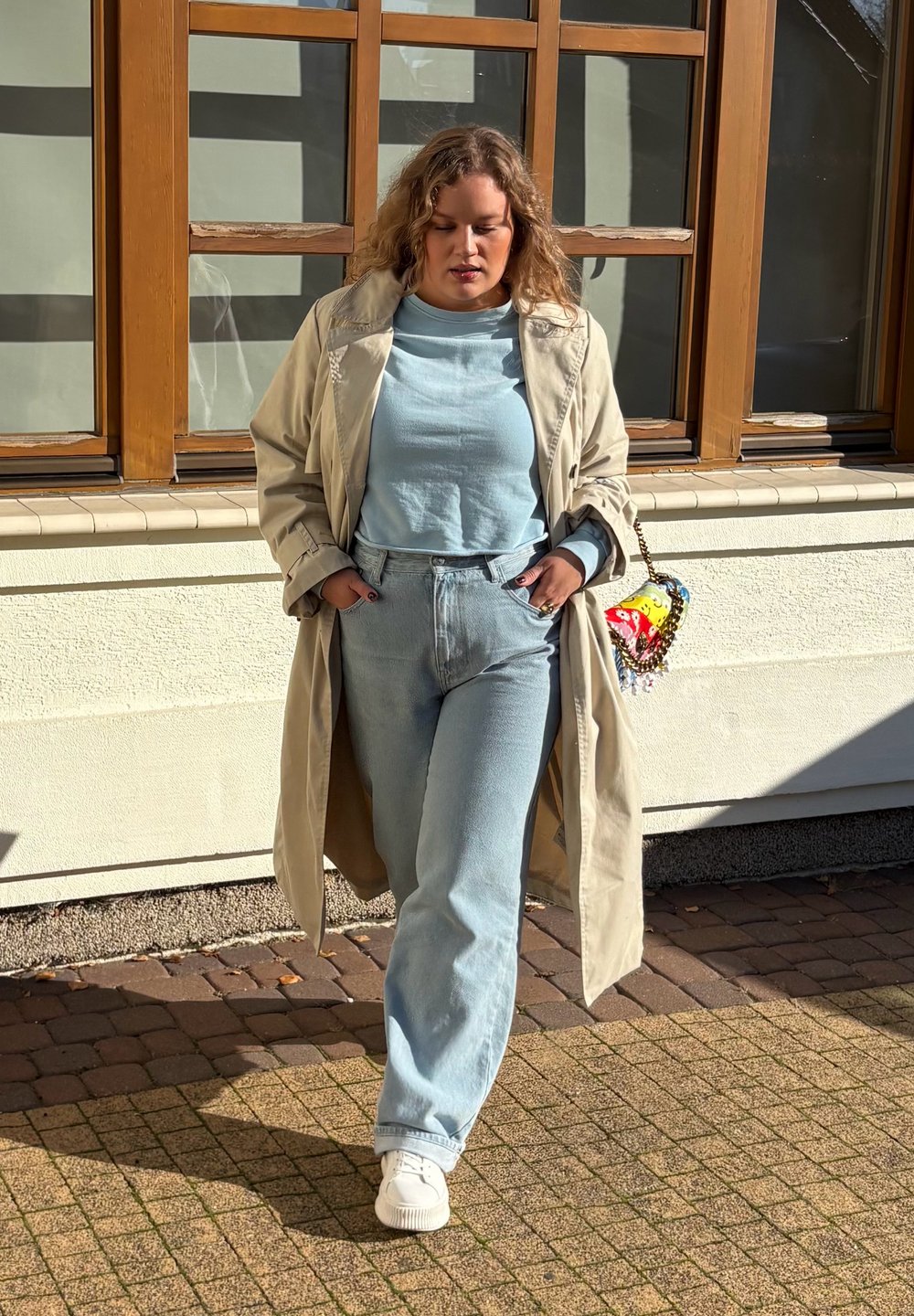 Bežový trenčkot přes světle modrý top a odpovídající džíny s vysokým pasem. Bílé tenisky. Malá vícebarevná kabelka s dekorativními detaily.