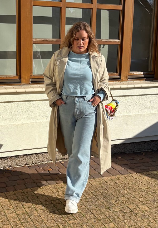 Beige trench coat egy világoskék felsőn és hozzáillő, magas derekú farmernadrágon. Fehér sportcipő. Kicsi, színes kézitáska díszítő részletekkel.