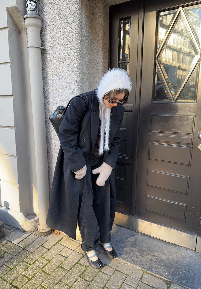 Langer schwarzer Mantel, helle, mit Fell gefütterte Kapuze, dunkle Jeanshose, beige Fäustlinge und schwarze Schuhe mit hellen Akzenten, stehend an einer Holztür.