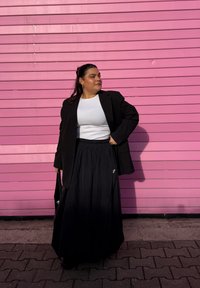 Saia longa preta com pregas, combinada com um top branco justo e um blazer preto oversized. O fundo apresenta uma parede rosa texturizada.