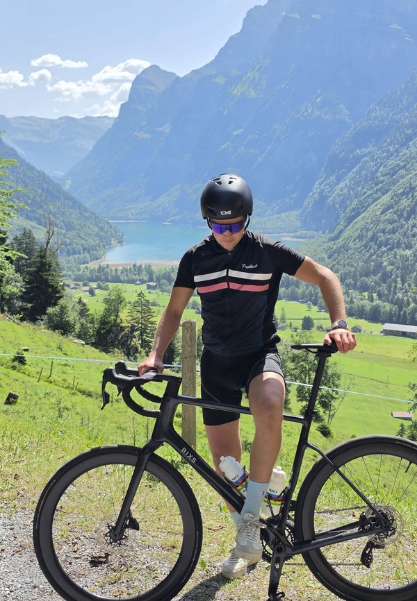 Fietser met helm en zonnebril staat met zwarte racefiets op een bergpad met uitzicht op een meer en een groene vallei.