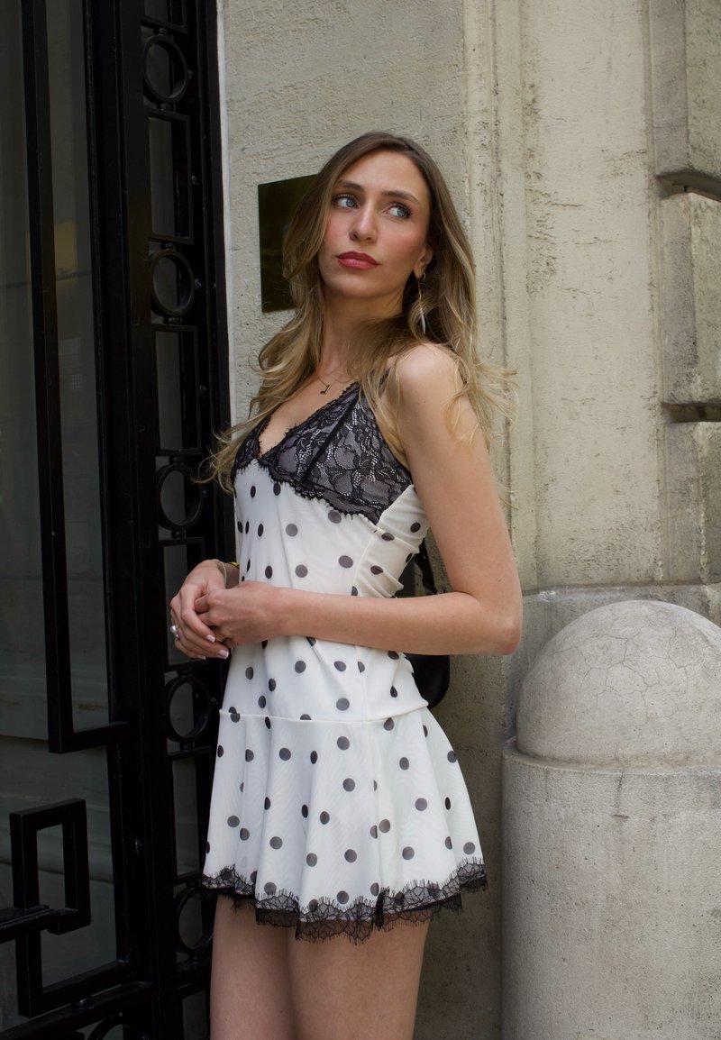 Jeune femme en robe blanche à pois avec des détails en dentelle noire, debout près d’un mur en pierre et d’un portail en métal noir, regardant sur le côté.