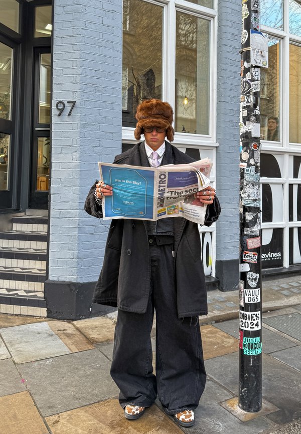 Person in oversized black coat and pants, wearing a brown fur hat and glasses, reading a newspaper on a city sidewalk near a sticker-covered pole.