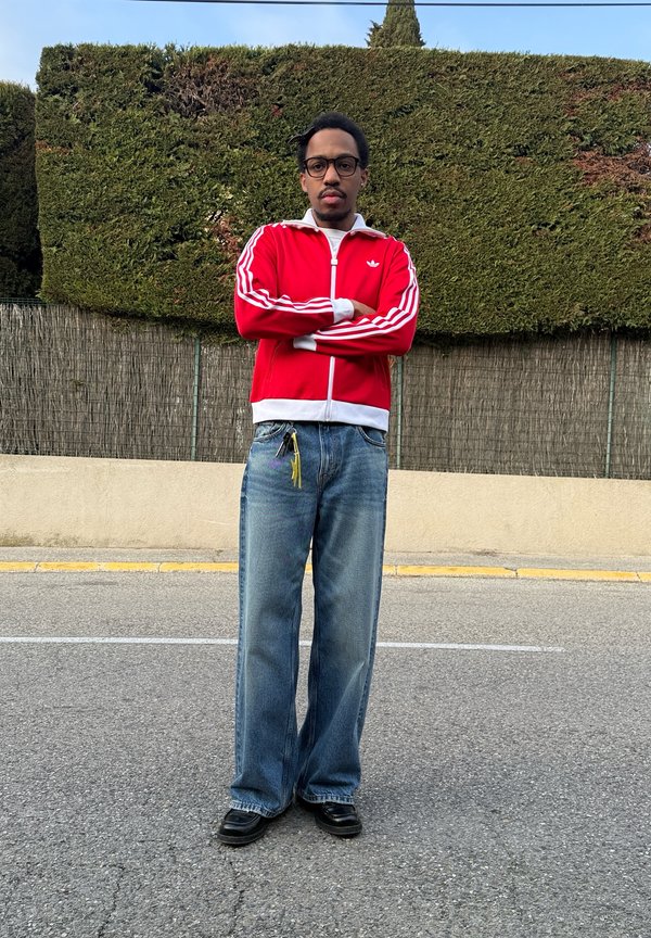 Mann med briller og bart som står på fortauet og har på seg rød Adidas-jakke, blå jeans, svarte sko, med armene i kors og grønn hekk i bakgrunnen.