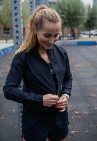 Chaqueta ligera negra con cremallera frontal, puños en las muñecas y un pequeño cuello, llevada sobre una camiseta negra y combinada con pantalones cortos. Entorno al aire libre.