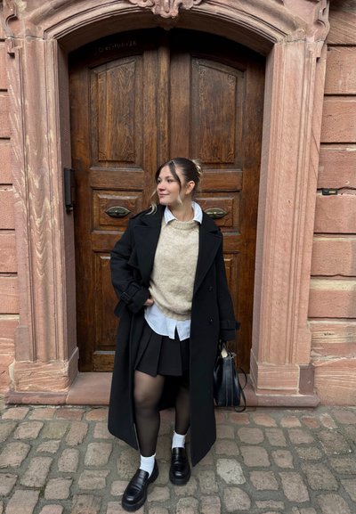 Mujer joven con abrigo negro, suéter beige, camisa blanca, falda negra y mocasines, de pie en una calle empedrada frente a una puerta de madera ornamentada y un arco de piedra.
