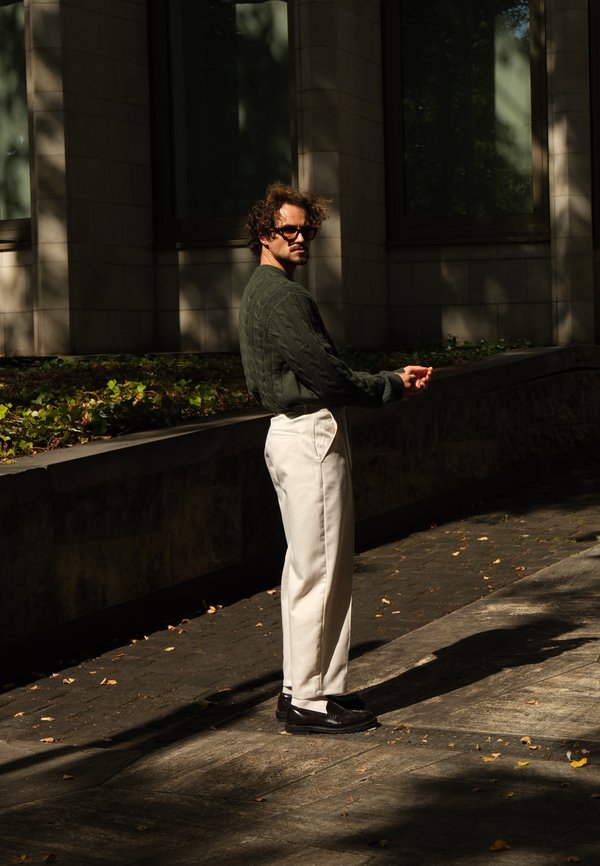 Grüner Pullover mit Zopfmuster, weiße maßgeschneiderte Hose, dunkle Schuhe. Mann steht im Freien, Sonnenlicht erzeugt Schatten auf der Steinoberfläche.