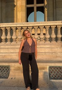 Ruskeat pilkulliset haalariyläosa, jossa edessä solmittava nauha, yhdistettynä mustiin leveälahkeisiin housuihin. Seisoo kivisestä balusterista vieressä lämpimässä auringonvalossa.