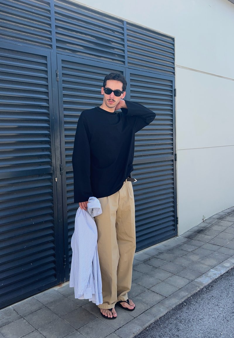 Homme portant une chemise noire à manches longues, un pantalon beige, des tongs et des lunettes de soleil, tenant une chemise rayée, debout près d'une porte métallique noire ventilée.