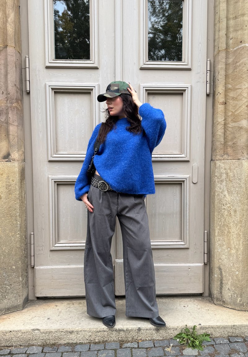 Blue fuzzy sweater, gray pinstripe wide-leg pants, black shoes, and a camouflage cap. Standing in front of light wooden double doors.
