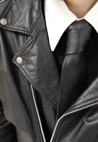 Veste de moto en cuir noir avec garnitures argentées, dotée d'une fermeture éclair diagonale, d'une finition texturée et d'un col superposé porté sur une chemise blanche.
