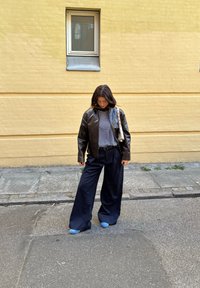 Chaqueta de cuero negra, camiseta de rayas, pantalones anchos negros y zapatos azul claro. Piel clara y cabello castaño, posando al aire libre frente a una pared amarilla.