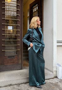 Grønnblå satengdress med vide bukser og en skreddersydd blazer, kombinert med en svart strukturert topp og spisse svarte sko, plassert foran en døråpning.