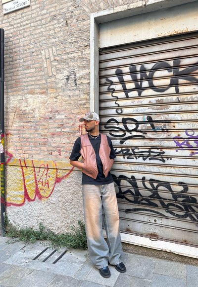 Chaleco de cuero marrón sobre una camisa negra, combinado con jeans de pierna ancha de color azul claro y zapatos negros. El fondo presenta una pared de ladrillo y graffiti.
