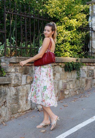 Vestido sin mangas de flores con patrones en rosa y verde, de tela ligera y diseño fluido. Bolso rojo y sandalias color beige tipo slip-on. Fondo de pared de piedra.