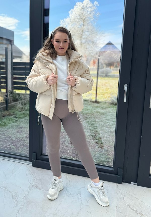 Casaco puffer creme com capuz, combinado com leggings caneladas bege e ténis brancos, contra um fundo interior iluminado.
