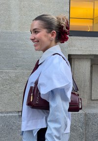 Borsa color marrone scuro con design strutturato, tasca con cerniera e scritta "EXPATRIATE". Indossata sopra una camicetta bianca con maniche voluminose e righe blu.