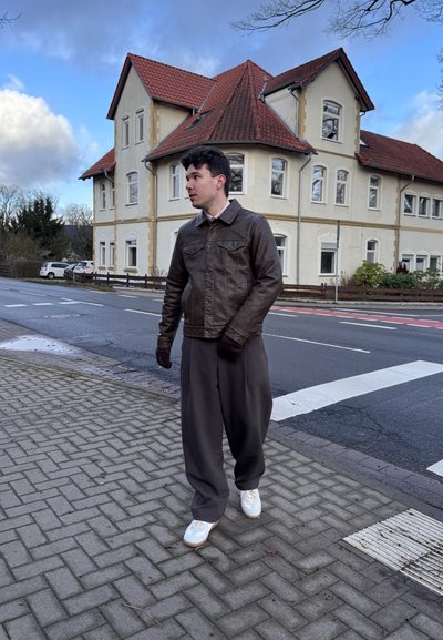 Chaqueta de cuero marrón, pantalones grises y zapatillas blancas. Caminando sobre un pavimento de ladrillos, con una casa de dos pisos al fondo.