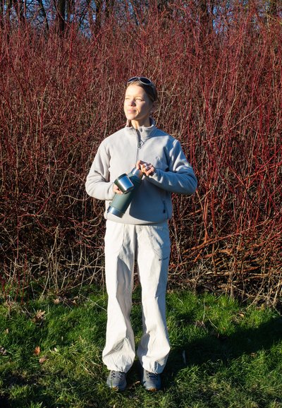 Chaqueta de forro polar gris, pantalones beige, sosteniendo un termo verde. Rodeado de ramas rojas y hierba verde, con luz solar brillante arriba.