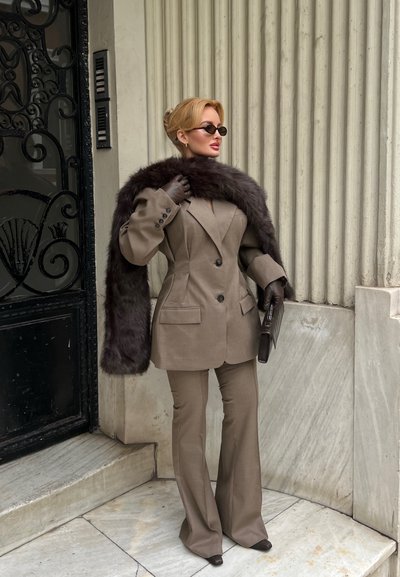 Mujer con traje topo, estola de piel negra, guantes y gafas de sol, de pie junto a una puerta negra ornamentada y columnas de piedra acanalada, sosteniendo un bolso de mano.