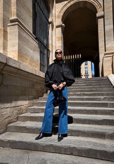 Blusa negra de manga abullonada con cuello con volantes, combinada con vaqueros de pierna ancha y botas negras hasta el tobillo, sosteniendo un pequeño bolso negro, en escalones de piedra.