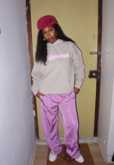 Sudadera gris claro con texto rosa, combinada con pantalones brillantes color lavanda. Calzado blanco y un gorro de piel rosa completan el atuendo.