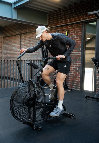 Hombre con gorra de béisbol y ropa deportiva pedaleando una bicicleta estática de aire en un gimnasio con paredes de ladrillo.