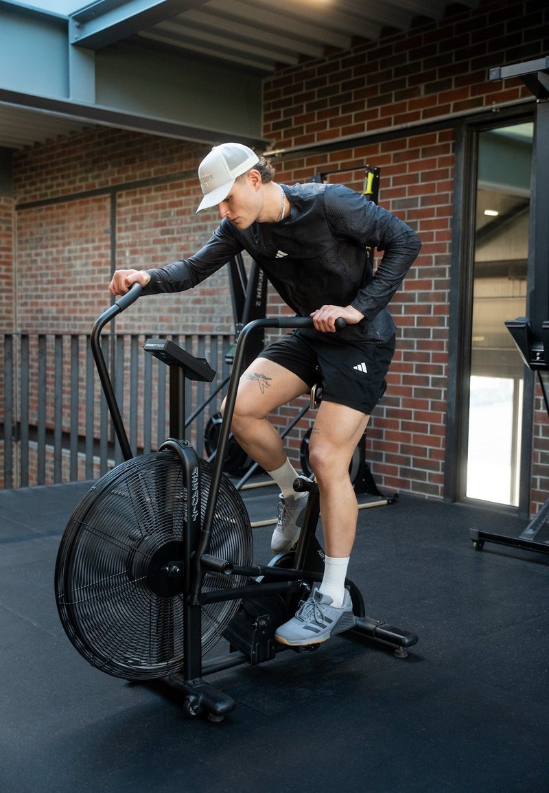 Man wearing a baseball cap and athletic wear pedals a stationary air bike in a gym with brick walls.