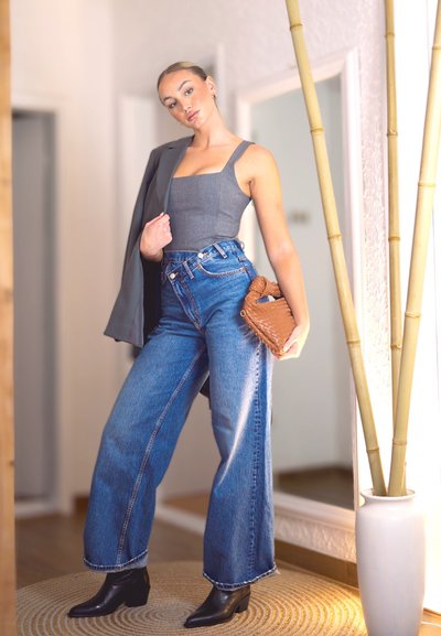 Top gris de cuello cuadrado, jeans azul de cintura alta, botines negros y un bolso de mano tejido marrón. Entorno interior con planta de bambú y espejos.
