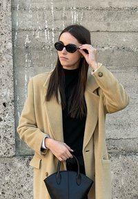 Beežist villast mantel suurte kraedega, must kõrge kaelusega pluus ja mustad ümmargused päikeseprillid, hoides väikest musta nahast käekotti. Tekstuurse betoonseina taust.