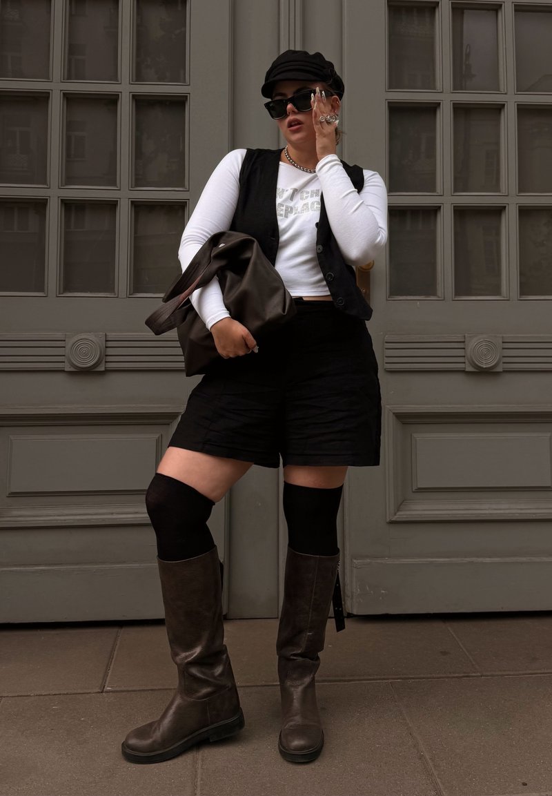 Colete preto sobre uma blusa de manga longa branca com texto prateado, combinado com calções pretos, botas castanhas até ao joelho e uma bolsa castanha. Boné preto e óculos de sol.