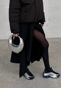 Casaco puffer preto, saia preta de fenda alta, collants pretos, ténis prateados e pretos, e uma bolsa prateada brilhante. Textura suave e design moderno.