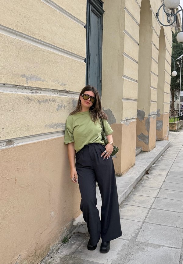 Mujer apoyada contra la pared beige de un edificio en la acera, vestida con camisa verde, pantalones anchos negros, botas negras, gafas de sol y llevando un bolso pequeño verde.