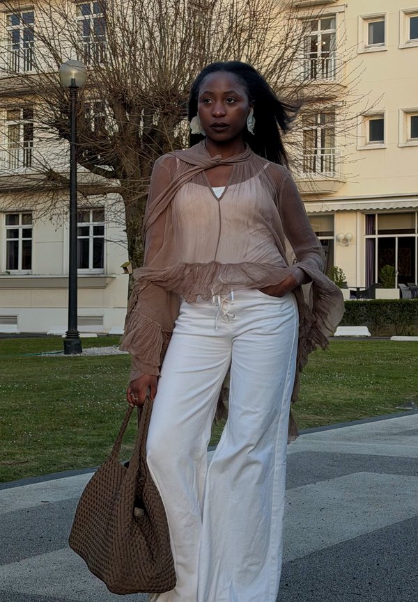 Femme portant un chemisier marron transparent et un pantalon blanc se tient dehors près d'un lampadaire, tenant un grand sac tressé marron.