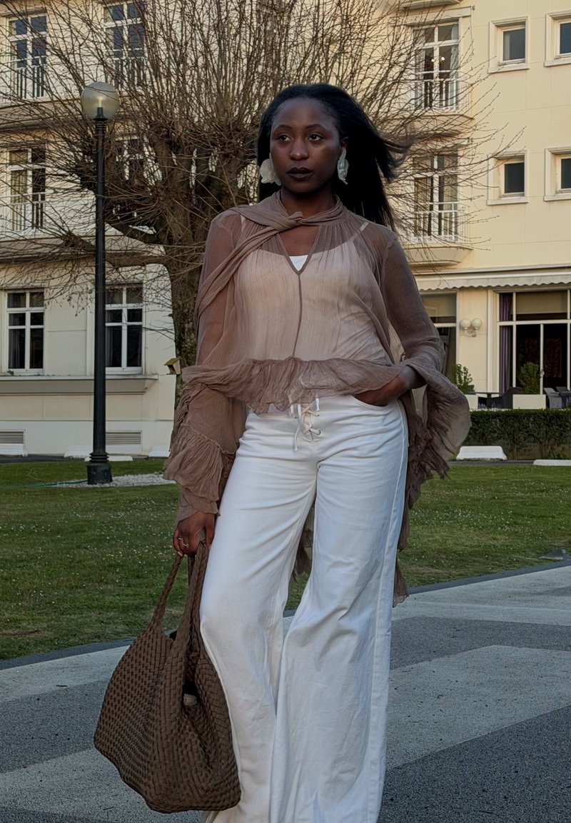 Vrouw met doorzichtige bruine blouse en witte broek staat buiten bij een lantaarnpaal, ze houdt een grote gevlochten bruine tas vast.