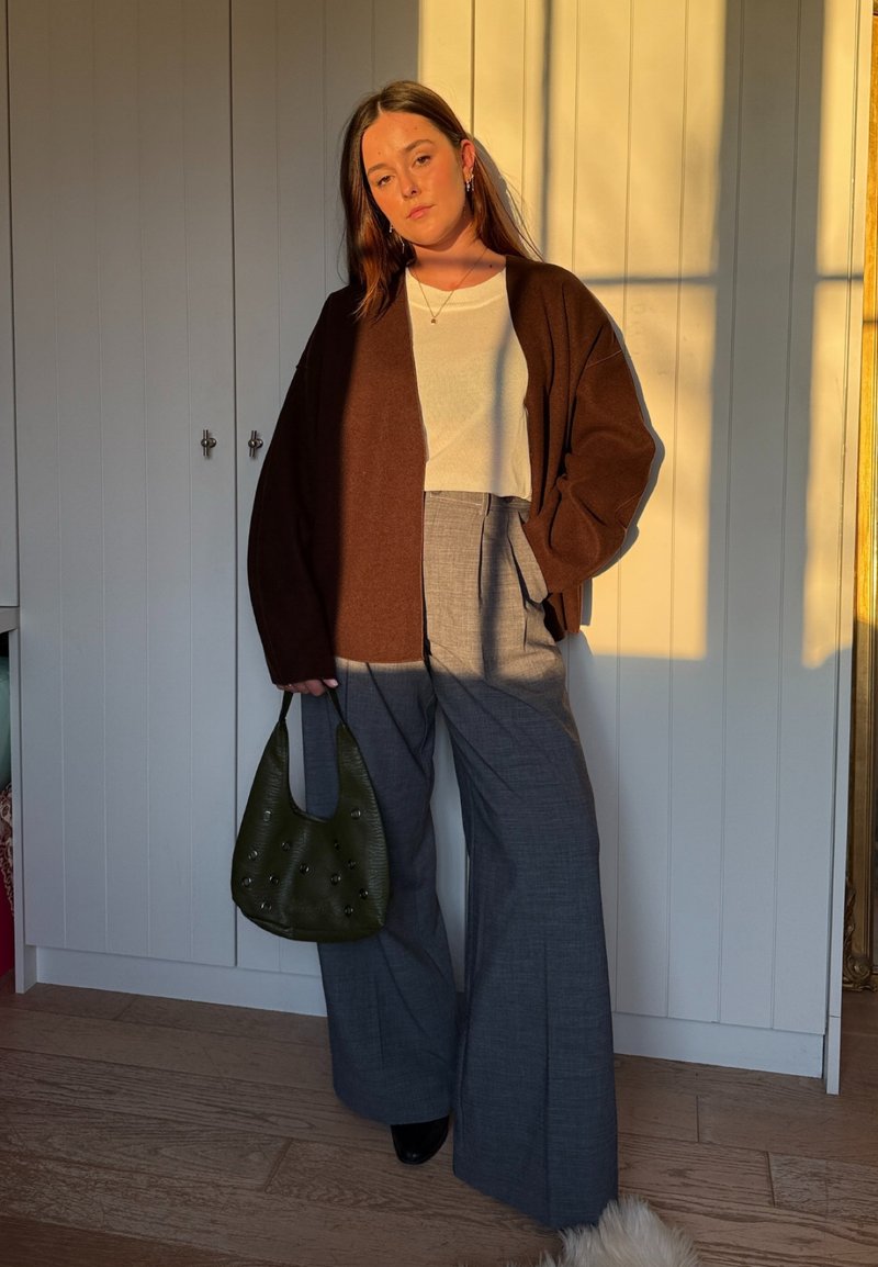 Femme debout à l'intérieur portant un cardigan marron, un haut blanc, un pantalon large gris, tenant un sac à main vert, avec la lumière du soleil projetant des ombres sur le mur derrière elle.