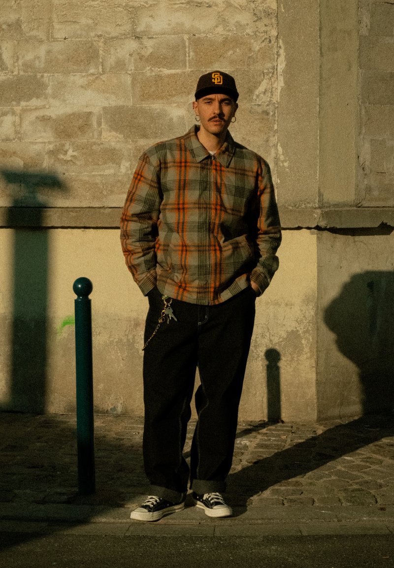 Homme portant une veste à carreaux, un pantalon noir avec des clés attachées, une casquette noire et des baskets, se tient devant un mur de pierre texturé au soleil.