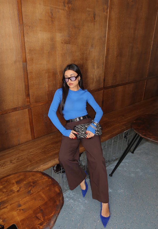 Une femme portant des lunettes de soleil, un haut bleu et un pantalon brun est assise sur un banc en bois et tient une pochette à imprimé zèbre dans une pièce lambrissée.