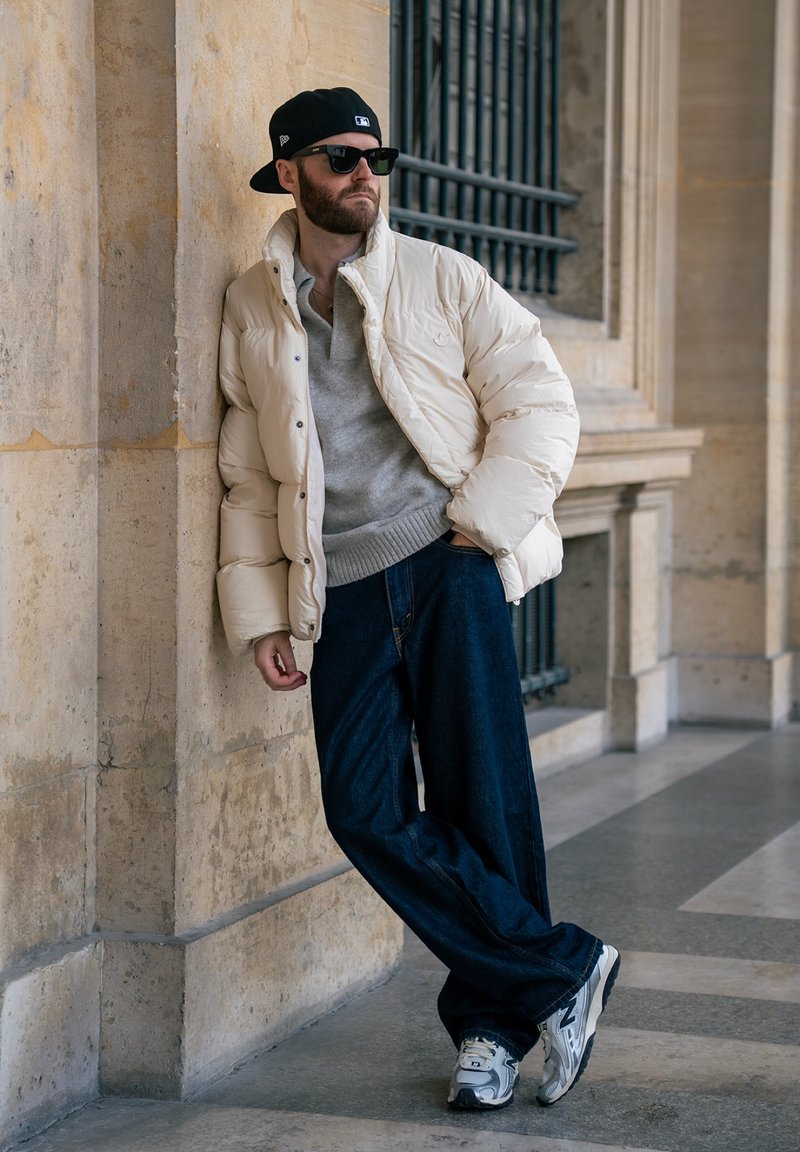 Homme barbu portant une casquette noire, des lunettes de soleil, une doudoune beige, un pull gris, un jean large foncé, des baskets grises, appuyé contre un mur en pierre à l'intérieur.
