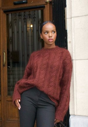 Suéter grueso de punto en burdeos profundo, con cuello redondo y corte suelto, combinado con pantalones negros ajustados. Fondo de puerta de madera.