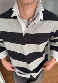 Homme portant un polo à rayures horizontales noir et gris par-dessus une chemise blanche à col avec manches retroussées, debout à l'intérieur.