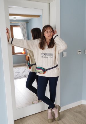 Mujer con suéter color crema, leggings azul marino y botas beige apoyada en un marco de puerta, con una bolsa de cintura colorida y un espejo reflectante detrás de ella.