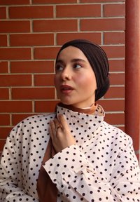 Joven mujer con un pañuelo negro y una chaqueta blanca con lunares, tocándose el cuello, mirando hacia un lado contra un fondo de pared de ladrillos.