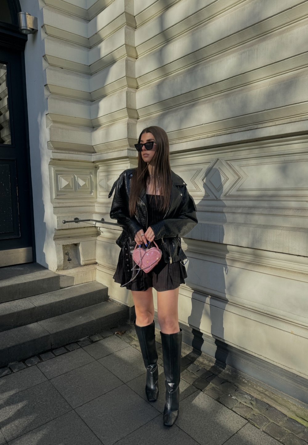 Svart läderjacka över en kort, plisserad svart klänning; knähöga svarta stövlar; håller en rosa hjärtformad väska; står mot en texturerad vägg.