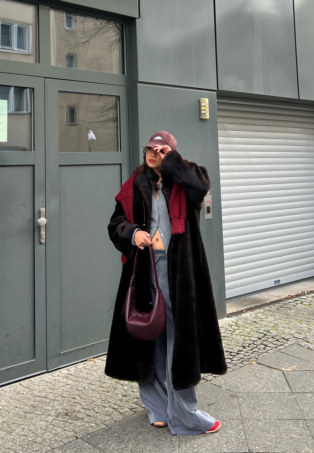 Čierny dlhý kabát s textúrovaným povrchom, bordový šál, sivý top, džínsové nohavice s širokými nohavicami a bordová kabelka. Žena s bordovou čiapkou.