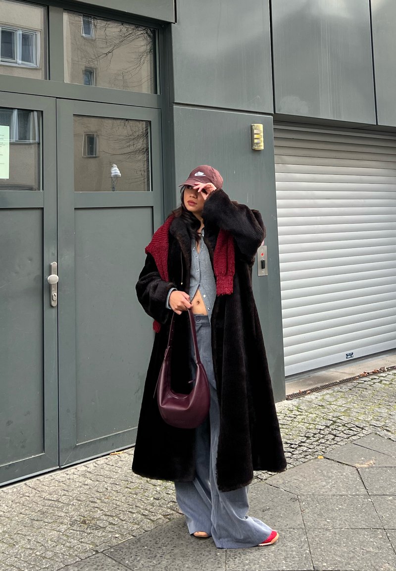 Cappotto lungo nero con finitura testurizzata, sciarpa bordeaux, top grigio, jeans a gamba larga e borsa bordeaux. Donna che indossa un berretto bordeaux.