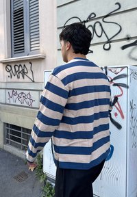 Csíkos, hosszú ujjú ing kék és bézs színben, puha pamut anyag, laza szabás, oldalsó hasítékokkal, fekete nadrágra viselve, graffitis háttér előtt.