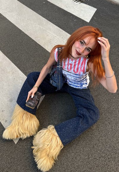 Peto vaquero con detalles de piel beige en los tobillos, combinado con una camiseta sin mangas blanca con un gráfico. Una persona sostiene un teléfono en una mano, sentada en un paso de peatones.