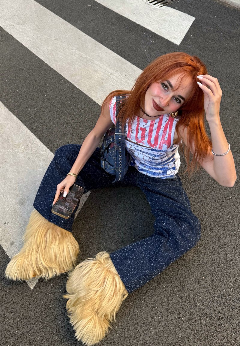 Salopette en denim avec des accents beiges en fourrure aux chevilles, assortie à un débardeur blanc à motif. Une main tient un téléphone, assis sur un passage piéton.